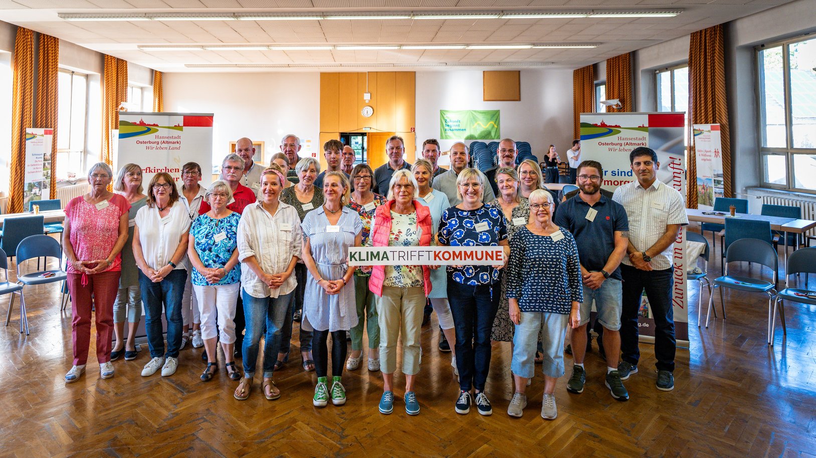 Das Bild zeigt die Mitglieder des Osterburger Bürgerrates bei ihrer Abschlusssitzung am 27. November 2025. Die Mitglieder stehen in drei Reihen; die erste Reihe hält ihre Teilnahmeurkunden in den Händen.  Im Hintergrund sind an der Seite jeweils Flipcharts zu sehen; in der Mitte wird eine Leinwand durch einen Beamer beleuchtet. Dort ist zu lesen: Osterburger Bürgerrat Mobilität.